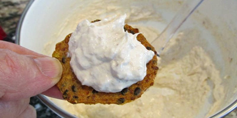 A hand holds a type of flat cracker topped with homemade smoked tuna cream cheese over a bowl of the same mixture.