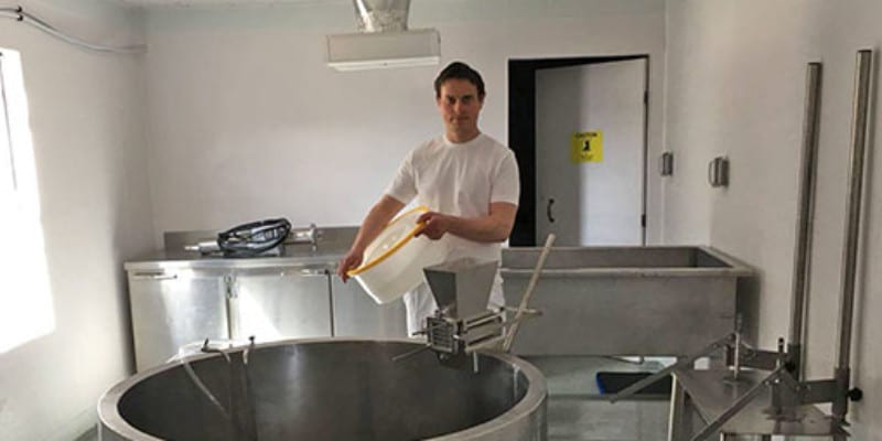 A young man in a white shirt and pants holds a large circular plastic container as he stands in a room used for cheesemaking. A large metal vat is in front of him with various metal cabinets and tubs are behind him.