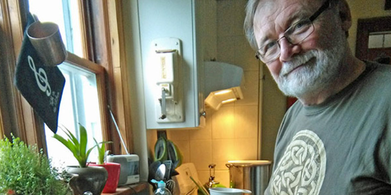 A white man with white facial hair and glasses wearing a dark green Ireland t-shirt stands at his kitchen sink. He is looking at the camera with a slight smile and slicing a loaf of homemade cheese on a white cutting board positioned on top of the sink basin. There is a window above the sink, and plants on the window sill. To his left, a white baking dish is filled with the slices of cheese he has already cut.