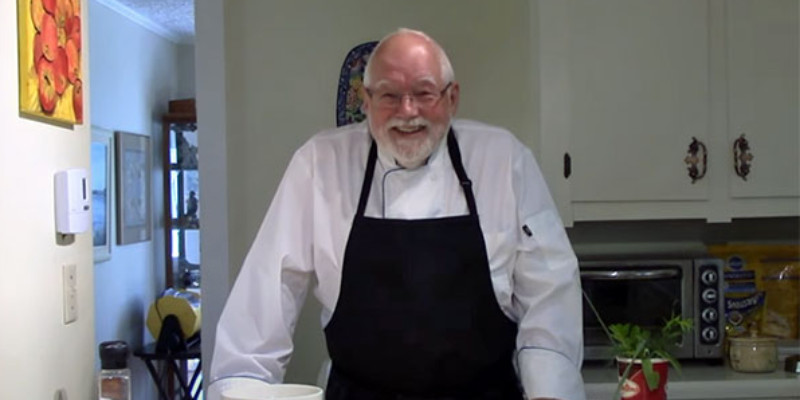 A white man with glasses, white receeding hair, and a white beard and mustache stands at a wooden countertop in a kitchen wearing a white chef's coat and black apron. There are kitchen cabinets behind him.