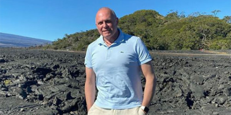 Cheesemaker Jamie Ryer, a white man with a short, military-style haircut stands on a hardened field of lava. He is wearing a light blue Lacoste polo, khaki shorts, and has his hands in his pockets.