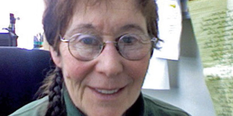 An older white woman with wire oval glasses, and brownish hair that is pulled back. A thin braid sits over her shoulder. She is wearing a dark green turtle neck and sitting in a black chair in front of a bulletin board with several white papers and a laminated green paper with writing on it.