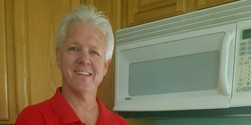 A white male with short white hair wearing a red polo shirt holds a wheel of cheese while standing in front of his stove. There is a stainless steel pot on the stove.