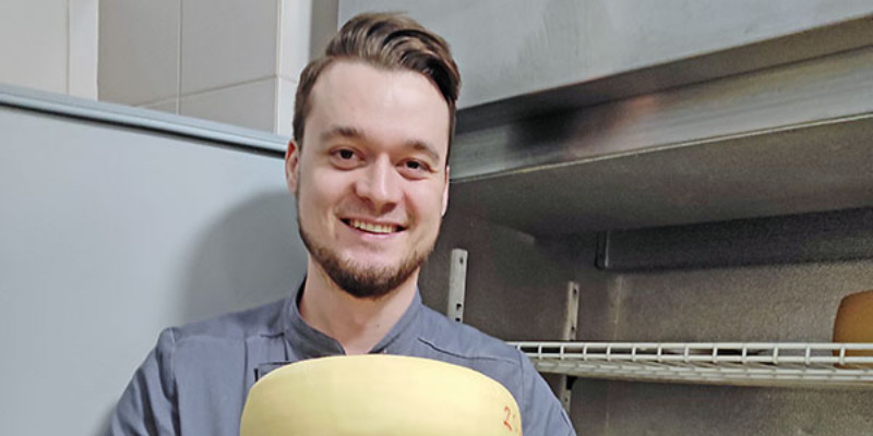 A younger white male holds five large wheels of cheese while smiling at the camera. He is wearing a gray shirt and black pants and is standing in front of what appears to be an open industrial refrigerator.