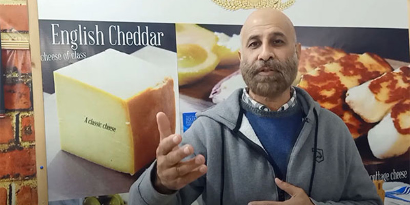 A screenshot of a YouTube video with a light-skinned man, bald, with a light brown beard, gray jacket over a blue sweater and collared shirt appears to be explaining something. There is a poster of cheese behind him.